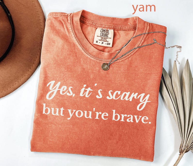 May include: A coral-colored Comfort Colors t-shirt with the text "Yes, it's scary but you're brave" in white. A silver necklace with a small pendant rests on the shirt. A brown hat and dried leaves are in the background.