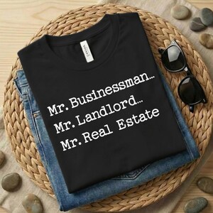 May include: Black t-shirt with white text: "Mr. Businessman... Mr. Landlord... Mr. Real Estate" laid on top of blue jeans. The shirt and jeans are on a woven, circular mat with sunglasses and small stones.