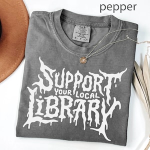 May include: A gray t-shirt with the text "SUPPORT YOUR LOCAL LIBRARY" in a white, stylized font. The shirt has a crew neck and short sleeves. A silver necklace with a small pendant is draped across the shirt.