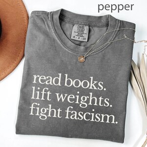 Könnte beinhalten: Graues T-Shirt mit dem Text "read books. lift weights. fight fascism." Eine silberne Halskette mit Anhänger liegt auf dem Shirt. Ein brauner Hut und eine getrocknete Pflanze befinden sich im Hintergrund.