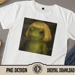 May include: White t-shirt featuring a square graphic of a green frog with a blonde bob haircut. The frog has large, expressive eyes. The shirt has a crew neck and short sleeves. The bottom of the shirt has a PNG design and digital download logo.