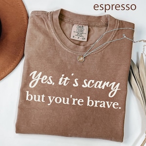 May include: Espresso-colored t-shirt with the text "Yes, it's scary but you're brave" in white. The shirt has a crew neck and a small tag. A silver necklace with a pendant is draped across the shirt.