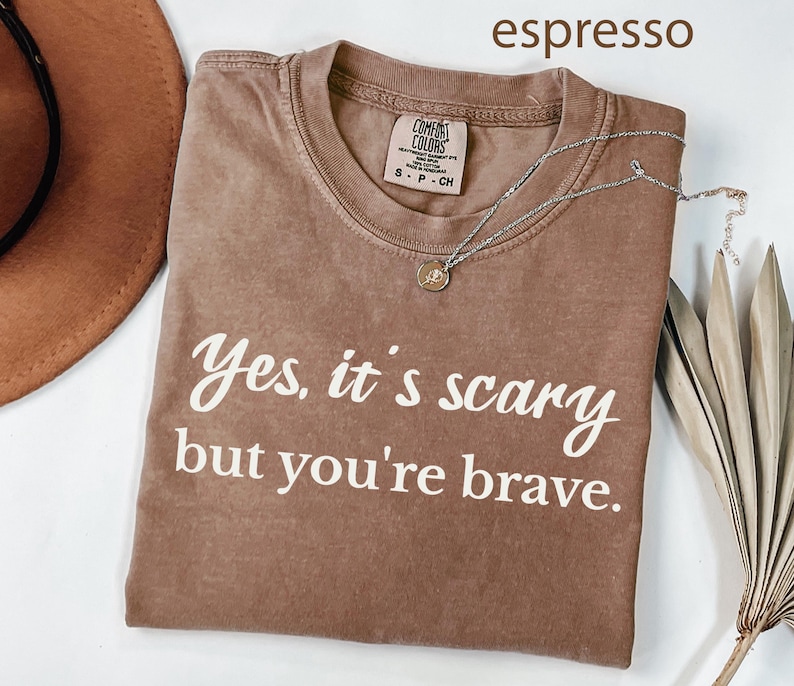 May include: Espresso-colored t-shirt with the text "Yes, it's scary but you're brave" in white. The shirt has a crew neck and a small tag. A silver necklace with a pendant is draped across the shirt.