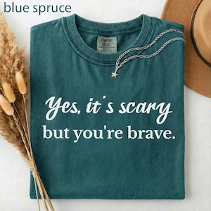 May include: A blue spruce t-shirt with the text "Yes, it's scary but you're brave" in white script. A silver necklace with a star pendant rests on the shirt. The shirt is folded and displayed with a hat and dried flowers.