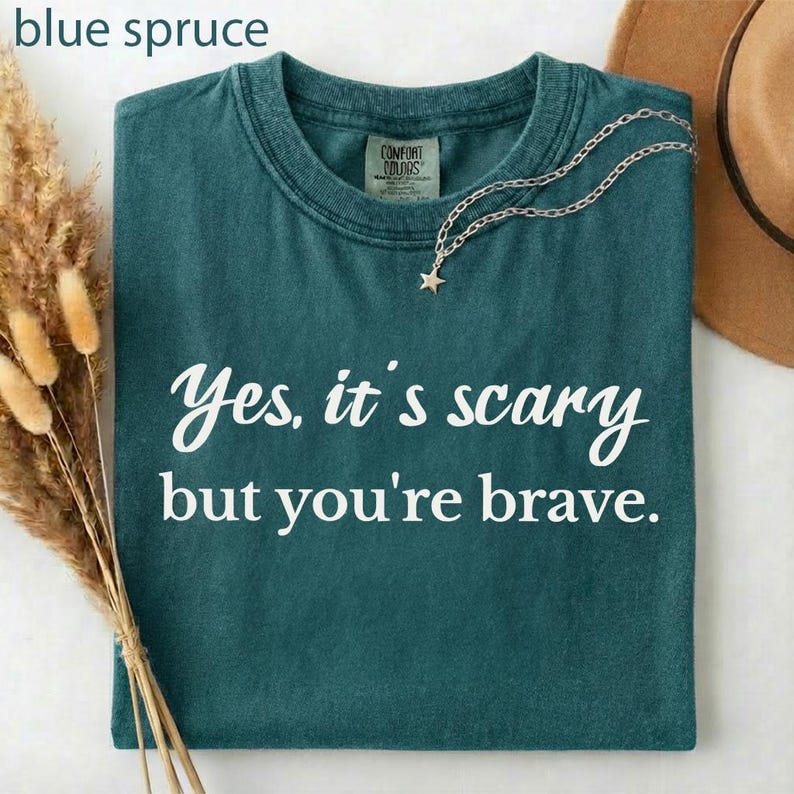 May include: A blue spruce t-shirt with the text "Yes, it's scary but you're brave" in white script. A silver necklace with a star pendant rests on the shirt. The shirt is folded and displayed with a hat and dried flowers.
