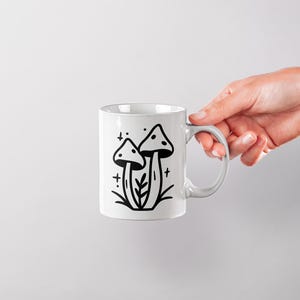 May include: White ceramic mug with a black line drawing of two mushrooms with a stem and cap. The mushrooms are surrounded by small stars and a few leaves.