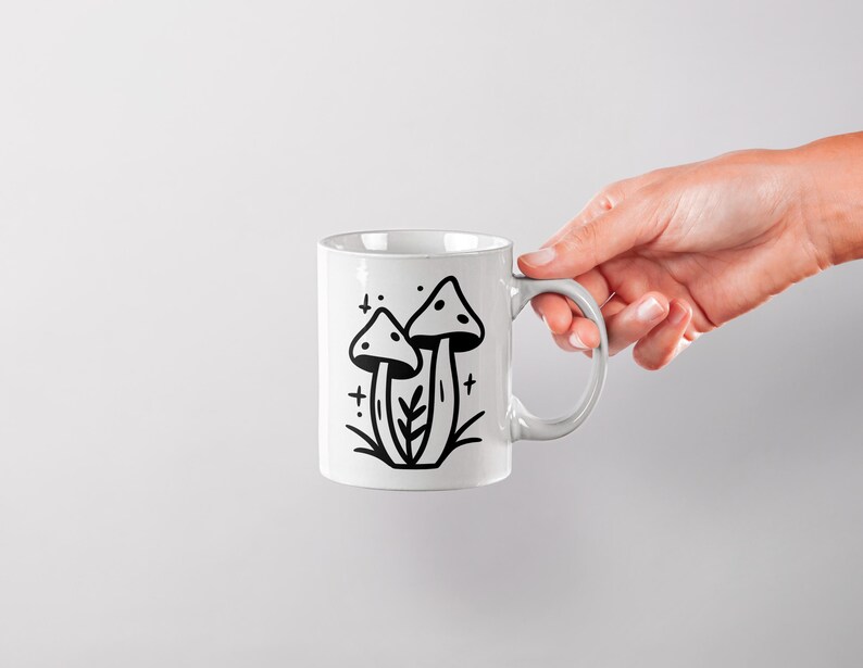 May include: White ceramic mug with a black line drawing of two mushrooms with a stem and cap. The mushrooms are surrounded by small stars and a few leaves.