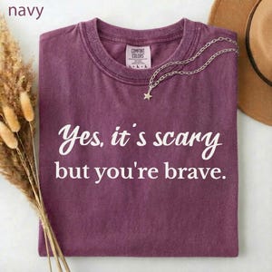 May include: A plum-colored t-shirt with the text "Yes, it's scary but you're brave" in white script. A silver necklace with a star pendant rests on the shirt. A brown hat is partially visible in the background.