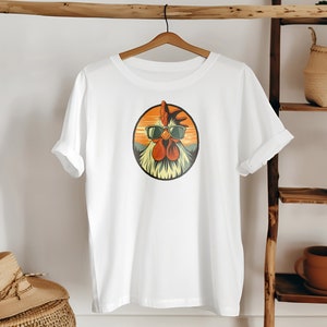 Op de afbeelding: Wit T-shirt met een afbeelding van een haan met een zonnebril in een cirkel met een oranje zonsondergang achtergrond.