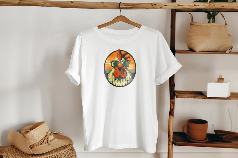 Op de afbeelding: Wit T-shirt met een afbeelding van een haan met een zonnebril in een cirkel met een oranje zonsondergang achtergrond.