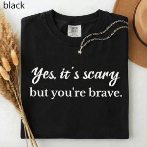 May include: Black t-shirt with the text "Yes, it's scary but you're brave" in white script. A silver necklace with a star pendant is draped on the shirt. A brown hat is visible in the background.