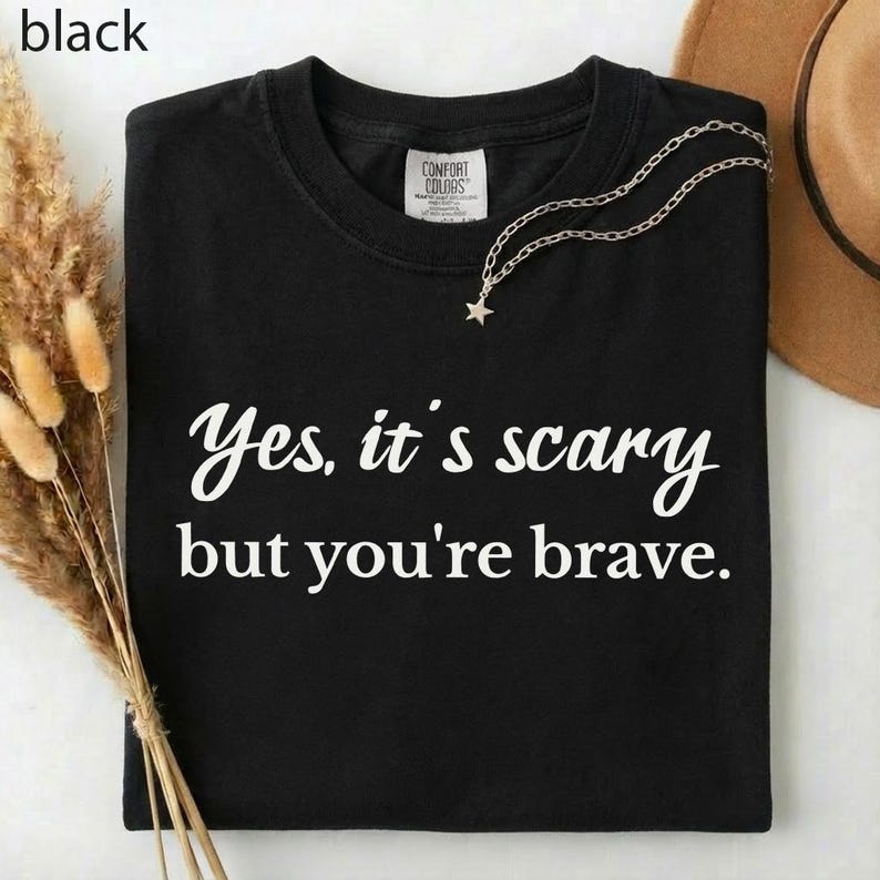 May include: Black t-shirt with the text "Yes, it's scary but you're brave" in white script. A silver necklace with a star pendant is draped on the shirt. A brown hat is visible in the background.