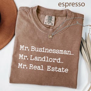 May include: Espresso-colored t-shirt with the text "Mr. Businessman... Mr. Landlord... Mr. Real Estate" in white. A silver necklace with a pendant rests on the shirt. The shirt is folded, with a brown hat and dried plant in the background.