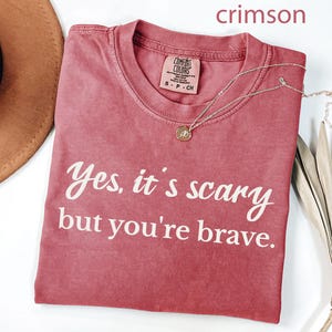 May include: Crimson-colored t-shirt with the text "Yes, it's scary but you're brave" in white script. The shirt has a crew neck and a small tag. A gold necklace with a pendant is draped across the shirt.