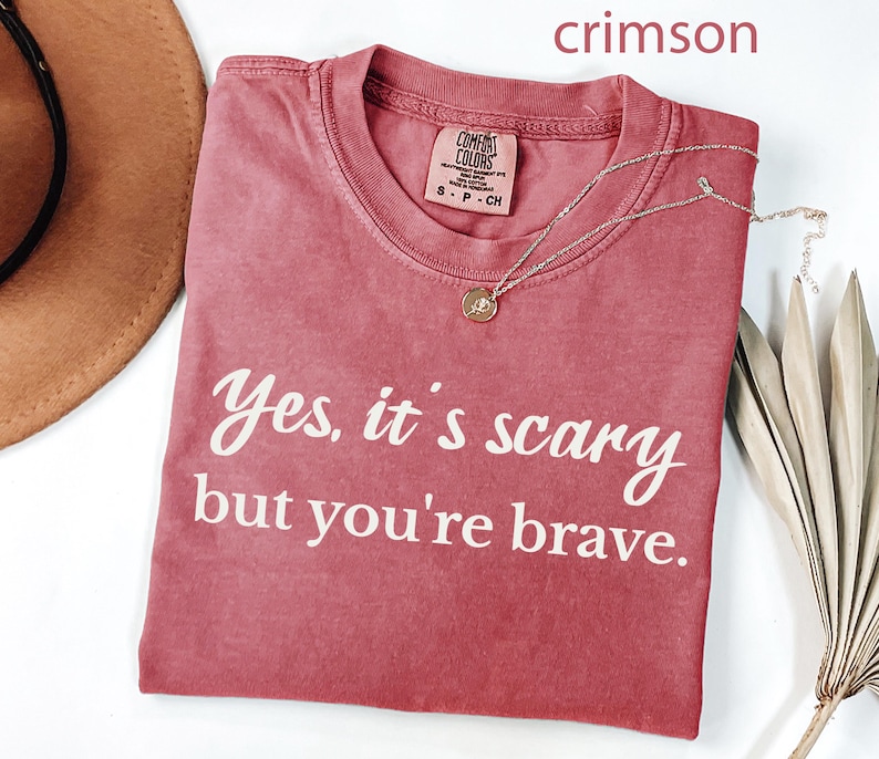 May include: Crimson-colored t-shirt with the text "Yes, it's scary but you're brave" in white script. The shirt has a crew neck and a small tag. A gold necklace with a pendant is draped across the shirt.