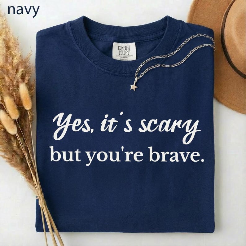 May include: Navy blue t-shirt with the text "Yes, it's scary but you're brave" in white script. A silver necklace with a star pendant rests on the shirt. The shirt is folded and displayed on a white surface.