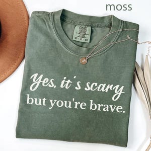 May include: A moss green t-shirt with the text "Yes, it's scary but you're brave" in white script. A gold necklace with a small pendant rests on the shirt. A brown hat and dried palm leaves are in the background.