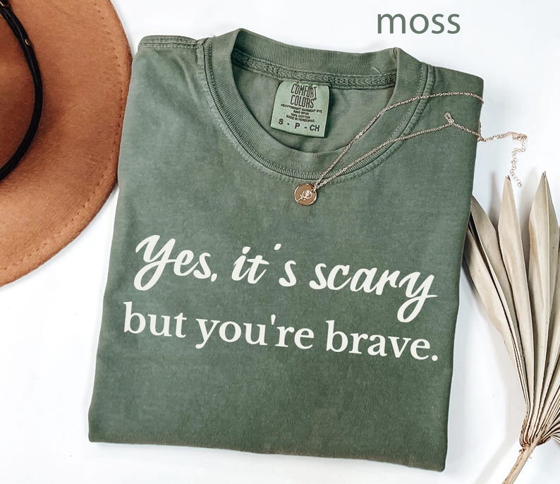 May include: A moss green t-shirt with the text "Yes, it's scary but you're brave" in white script. A gold necklace with a small pendant rests on the shirt. A brown hat and dried palm leaves are in the background.