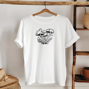 May include: White t-shirt with a black and white illustration of three mushrooms with a dotted pattern on the tops. The mushrooms are surrounded by leaves and stars.