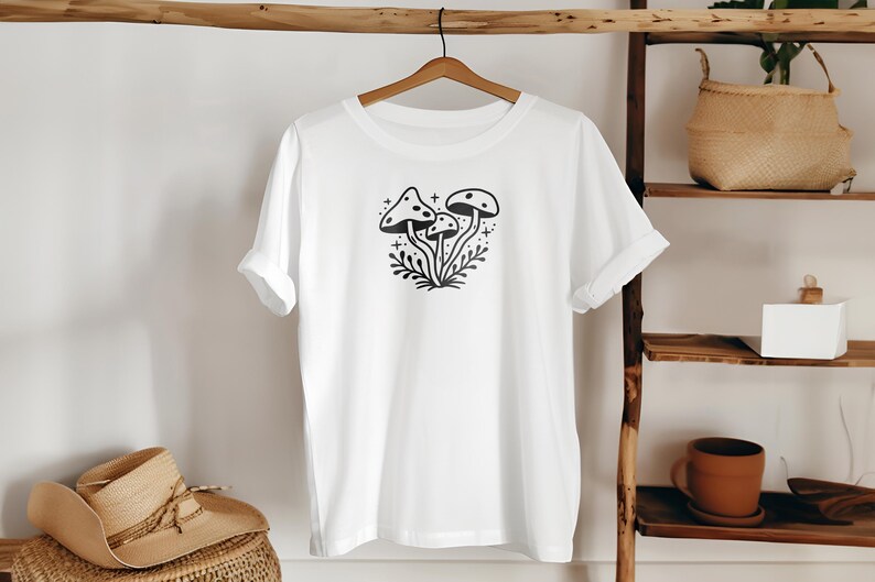 May include: White t-shirt with a black and white illustration of three mushrooms with a dotted pattern on the tops. The mushrooms are surrounded by leaves and stars.