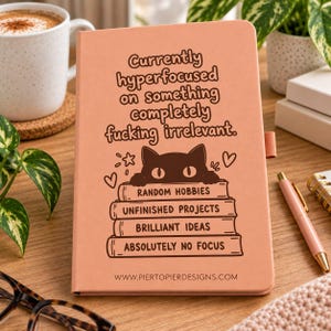 May include: A peach-coloured notebook featuring a dark brown illustration of a cat peeking over a stack of four books labelled Random Hobbies, Unfinished Projects, Brilliant Ideas, and Absolutely No Focus, with text above reading Currently hyperfocused on something completely fucking irrelevant.
