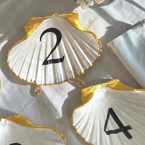 Puede incluir: Porta números de mesa con forma de concha blanca con borde dorado y números negros. Los números 2 y 4 son visibles. Los soportes tienen pequeños soportes dorados. Ideal para bodas o eventos.