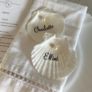 May include: Two white seashell place cards, each with a pearl accent. One shell reads "Charlotte" and the other reads "Elliot" in black script. The place cards rest on a white linen napkin, with a menu visible in the background.