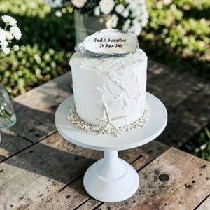 May include: A white frosted cake with a textured finish on a white pedestal stand. The cake is topped with a silver-rimmed oyster shell featuring the text Paul & Jacqueline 24 June 20XX. A single starfish and small white seashells decorate the base.