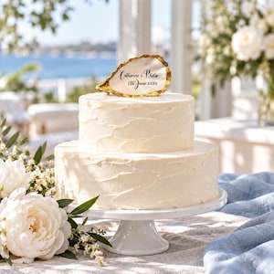 May include: A two-tier white frosted cake on a white pedestal stand, topped with a gold-rimmed oyster shell featuring the black printed text Catherine & Philip 11th June xxxx. The cake has a textured finish and sits near white flowers.