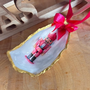 Könnte beinhalten: Weihnachtsschmuck mit einer bemalten Nussknackerfigur in einer weißen Austernschale mit goldenem Rand. Oben ist eine leuchtend rosa Schleife gebunden. Der Schmuck liegt auf einer Holzoberfläche.