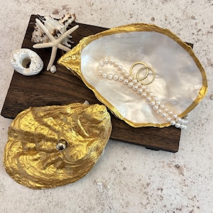 May include: A decorative oyster shell trinket dish with a gold-painted exterior and a pearlescent white interior. The dish contains a strand of white pearls and two gold rings. A separate gold-painted shell lid with a small knob sits nearby.