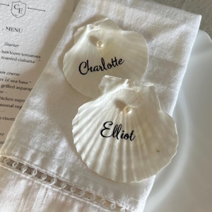 Könnte beinhalten: Zwei weiße Jakobsmuscheln als Platzkarten, jeweils mit einer kleinen weißen Perle an der Oberseite und einem in schwarzer Schreibschrift aufgedruckten Namen. Eine Muschel ist mit dem Namen Charlotte und die andere mit Elliot beschriftet.