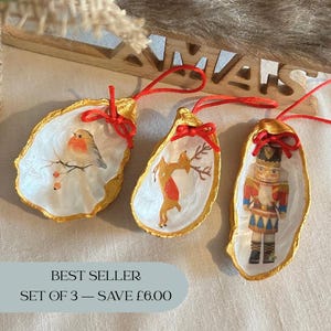 May include: Three oyster shell Christmas ornaments with red hanging ribbons. Each shell has a gold rim and features a different hand-painted design: a robin, a deer, and a nutcracker. The text "BEST SELLER" and "SET OF 3 - SAVE £6.00" is visible.