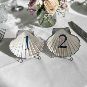May include: Two seashell table number holders. The shells are white with silver edging and feature the numbers 1 and 2 in teal. Each shell sits on a small, decorative metal stand. A vase of flowers is in the background.
