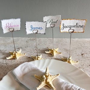 May include: Four place card holders featuring cream-colored starfish bases and thin silver wire stems with heart-shaped clips. Each holds a rectangular piece of textured white paper with the names Rebecca, James, Paul, and Jacqueline printed.