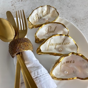 May include: Four oyster shells with gold-painted edges, each featuring a white pearl and a name written in gold script: Richard, Paul, Jacqueline, and James. A white napkin is held by a gold beaded napkin ring with gold cutlery.