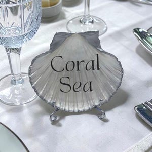 May include: A seashell-shaped place card holder with the words "Coral Sea" in black script. The holder is silver and white, resting on a silver wire stand. The background includes a white tablecloth and glassware.