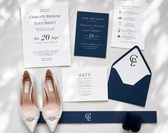May include: A wedding stationery set featuring white and navy blue cards with black text, including an invitation, save the date, details card, and RSVP card. A navy blue envelope with a monogram, a navy blue band, and white pointed-toe heels.