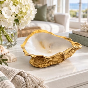 May include: A decorative oyster shell bowl featuring a textured, metallic gold exterior and a smooth, glossy white interior. A single small, round white pearl is positioned in the centre of the shell bowl, which rests on a flat surface.