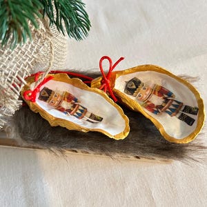 Könnte beinhalten: Zwei Weihnachtsschmuckstücke aus Austernschalen, jedes mit einem farbenfrohen Nussknacker-Design. Die Schalen sind an den Rändern goldfarben bemalt und haben eine rote Schnur zum Aufhängen. Der Schmuck liegt auf einer fellartigen Oberfläche.