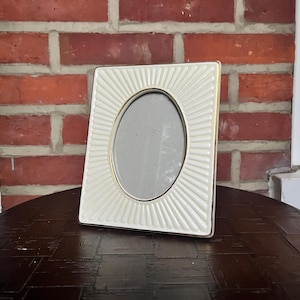 Puede incluir: Un marco de fotos rectangular con un borde de color crema que presenta un patrón de rayos de sol con líneas en relieve. El marco tiene una abertura central de forma ovalada y un fino borde metálico dorado alrededor del área interior.