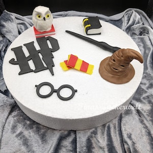 Könnte beinhalten: Eine weiße Fondant-Eule, ein schwarzer Fondant-Zauberstab, ein rot-gelber Fondant-Schal, eine schwarze Fondant-Brille, ein schwarzes Fondant-'HP'-Logo mit einem Blitz, ein brauner Fondant-Hexenhut und ein schwarzes Fondant-Buch auf einem weißen Kuchenbrett.