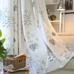 May include: White sheer curtains with a floral pattern of green leaves and white flowers. The curtains are hanging in a living room with a sofa and a lamp.