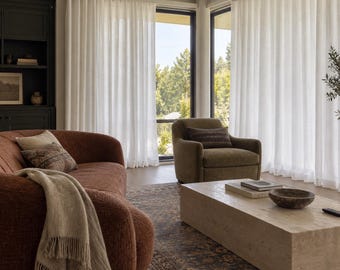 Può includere: Un soggiorno con un divano curvo color ruggine con una coperta beige, una poltrona abbinata e un tavolino rettangolare in travertino. Grandi finestre sono coperte da tende bianche trasparenti dal soffitto al pavimento.