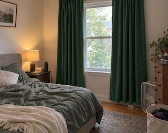 May include: A pair of floor-length, deep forest green curtains hanging on a dark rod over a window. The curtains feature a pleated header and a solid, matte texture, providing full coverage for the window frame in a bedroom setting.