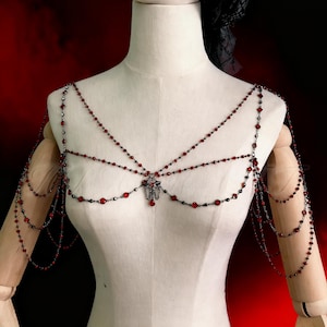 May include: A body chain necklace featuring red beads and a dark metal finish. The necklace is draped across the shoulders and chest, with multiple strands and a central pendant. The jewellery is displayed on a mannequin.