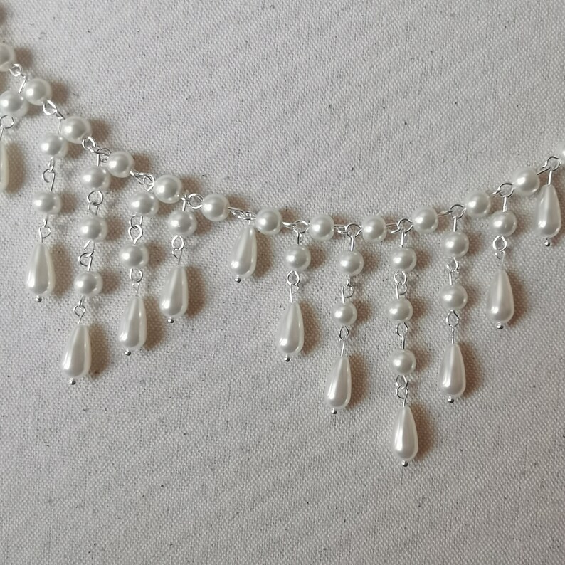 Op de afbeelding: Een witte parelketting met een cascade-ontwerp. De ketting heeft kleine ronde parels en grotere traanvormige parels.