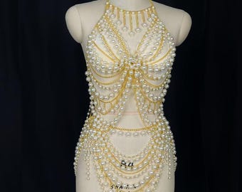 May include: A gold chain body harness with white pearls. The harness is made of multiple layers of chains and pearls, and it is designed to be worn over a bra or top.