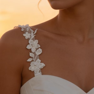 May include: A close-up of a white wedding dress with floral lace straps. The straps are adorned with delicate, three-dimensional flowers. The background is a warm, golden sunset, creating a soft, romantic ambiance. The dress appears to be made of a smooth, satin-like fabric.