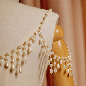 Op de afbeelding: Een gouden ketting met witte parels en hangende pareldruppels. De ketting is over een wit stoffen mannequin gelegd.